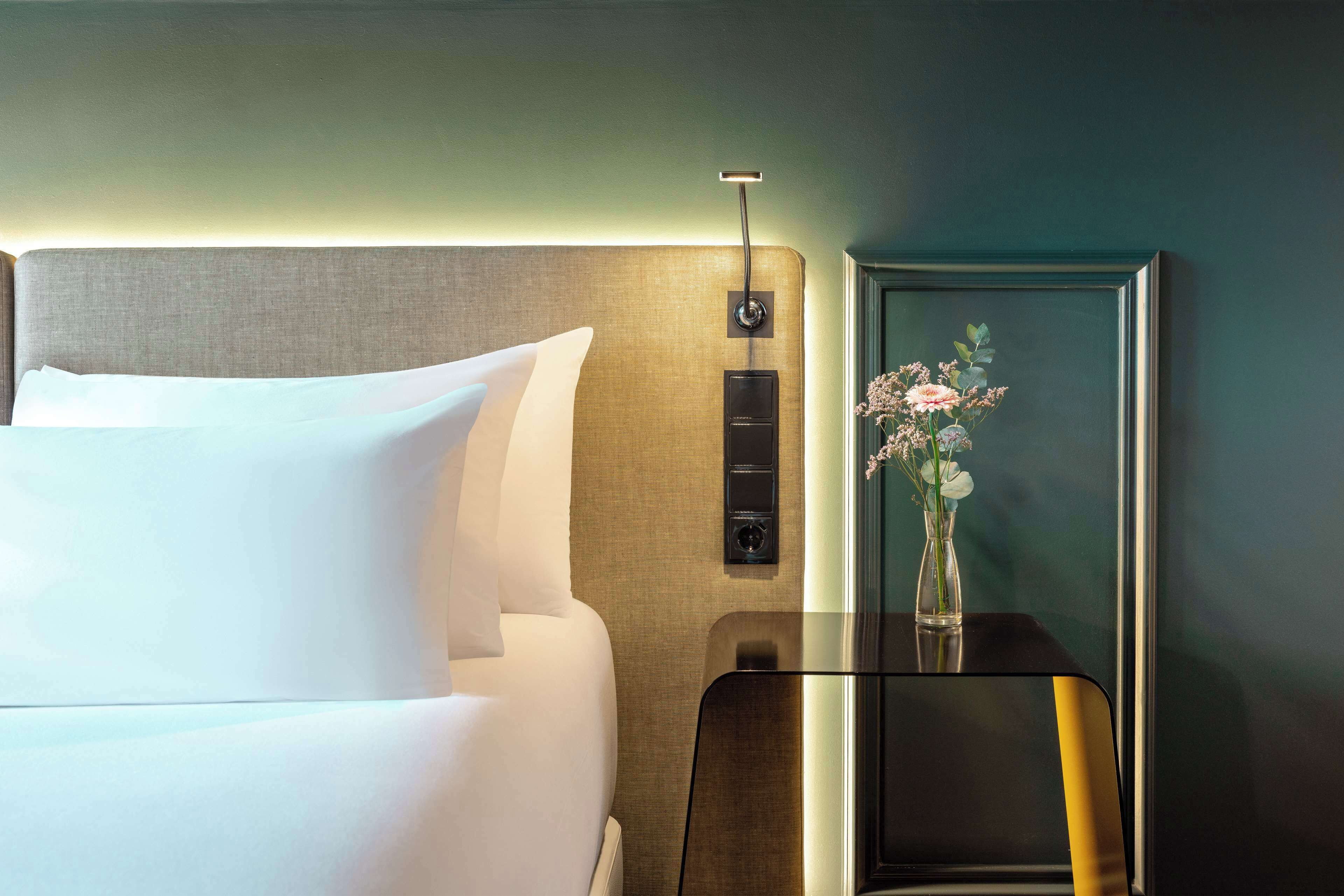 Habitación de hotel con una cama bien hecha, una mesita de noche con un jarrón de flores y un panel de control en la pared.