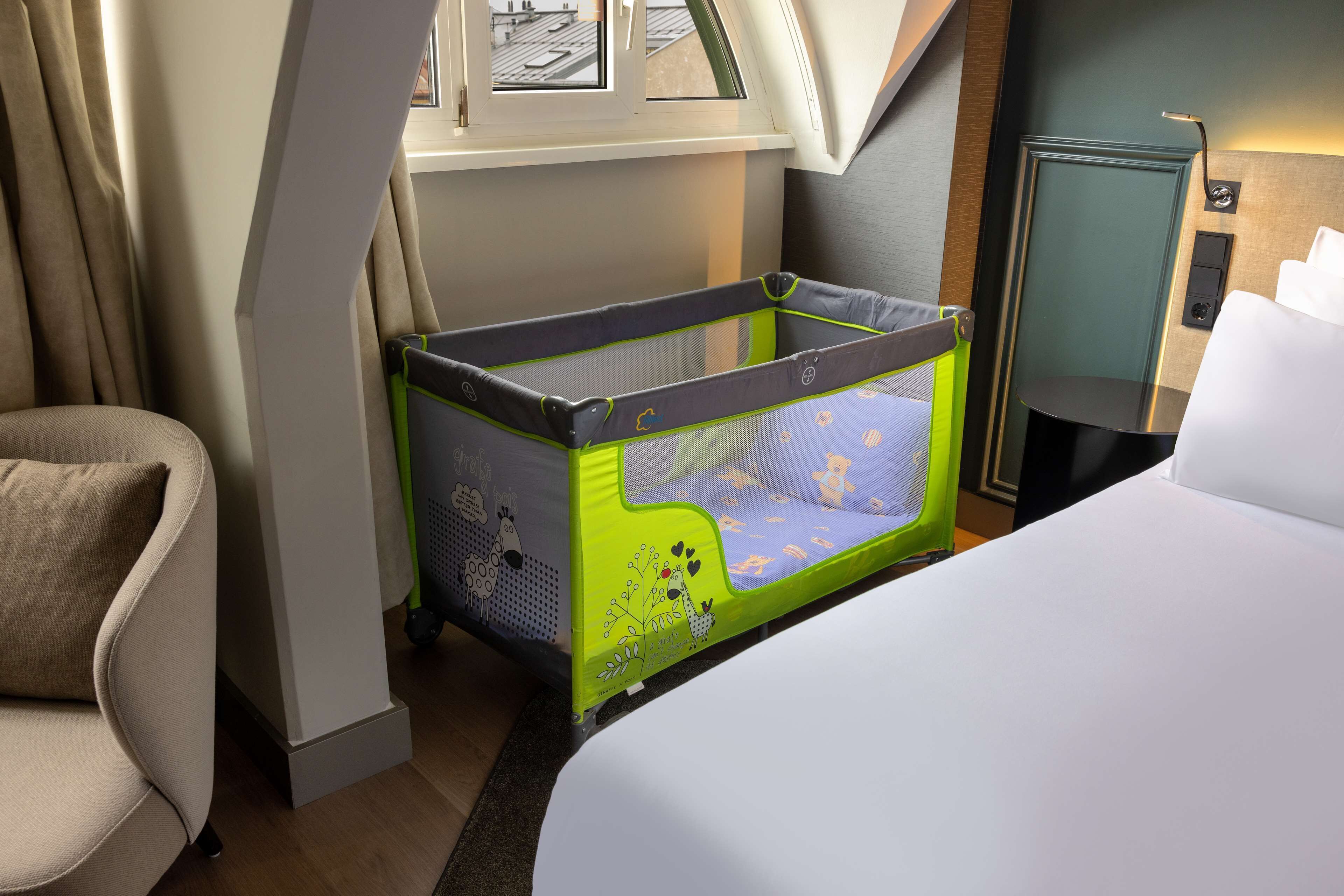 A hotel room with a bed, a green and blue baby crib, a window with curtains, and a view of the cityscape.