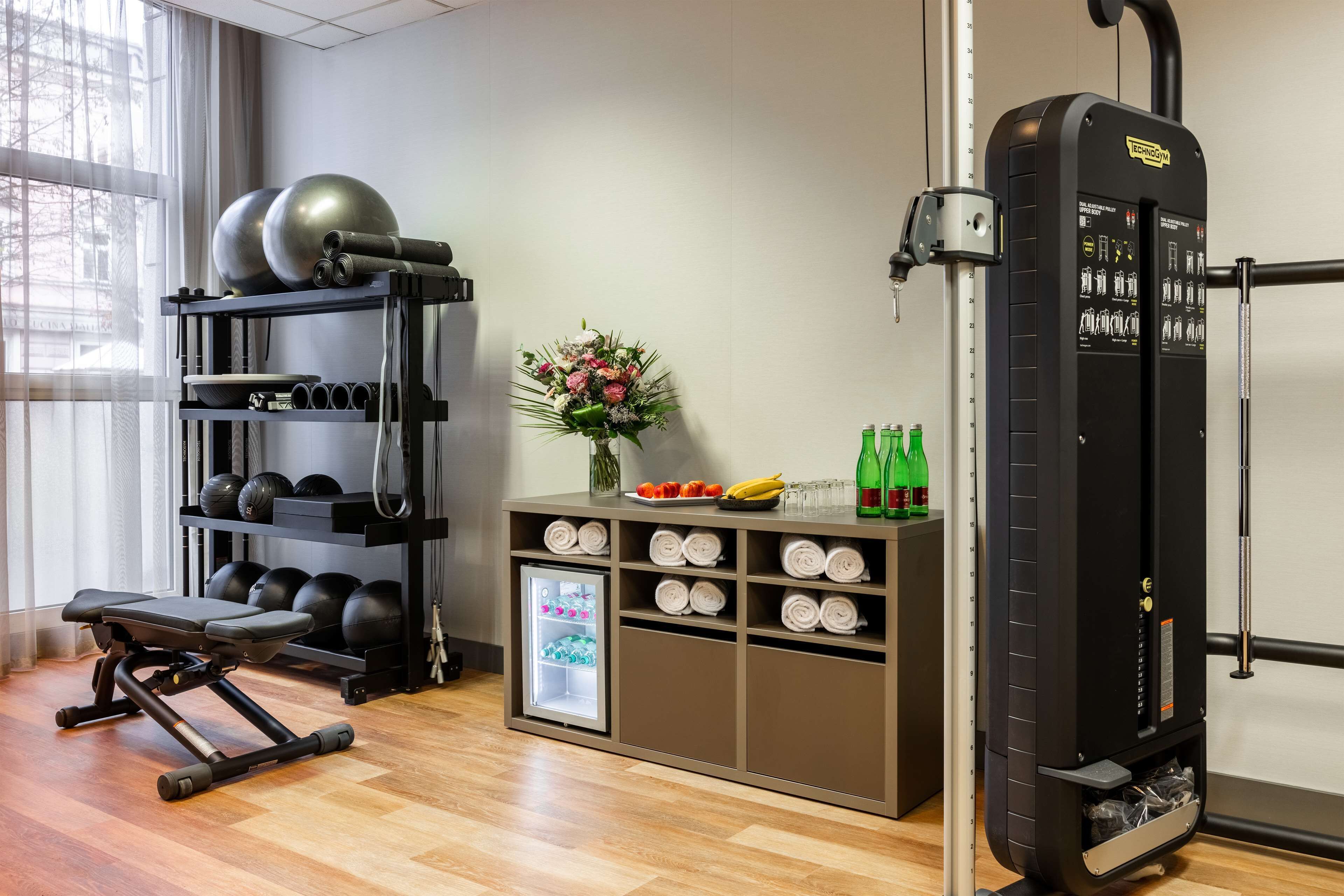 Un gimnasio con equipamiento de ejercicio, una banca, un estante con pesas en la pared, un refrigerador pequeño con bocadillos y un arreglo floral.