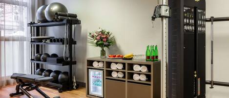 Un gimnasio con equipamiento de ejercicio, una banca, un estante con pesas en la pared, un refrigerador pequeño con bocadillos y un arreglo floral.
