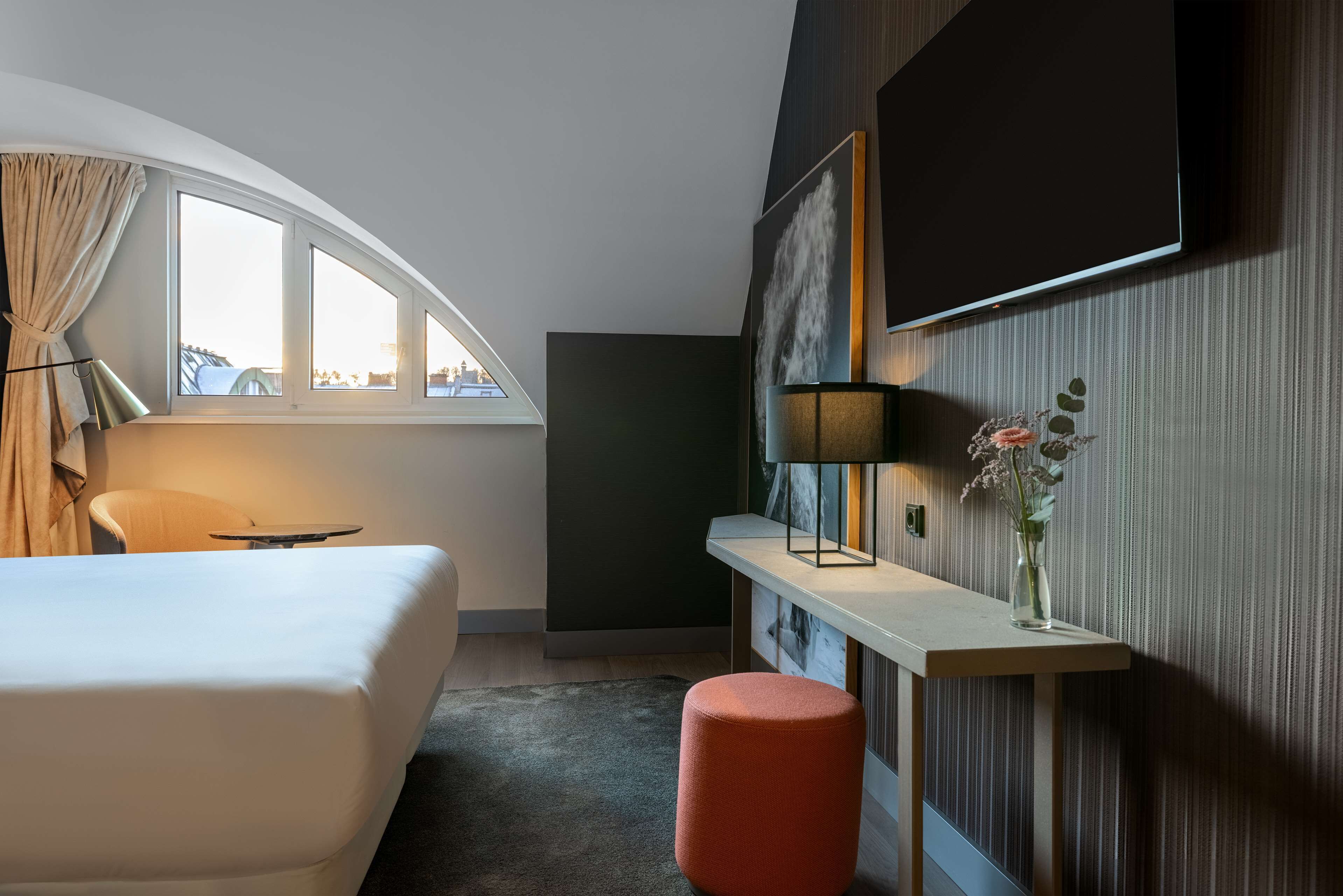 Una habitación de hotel moderna con cama, escritorio, lámpara, un florero con flores y un amplio ventanal con cortinas.