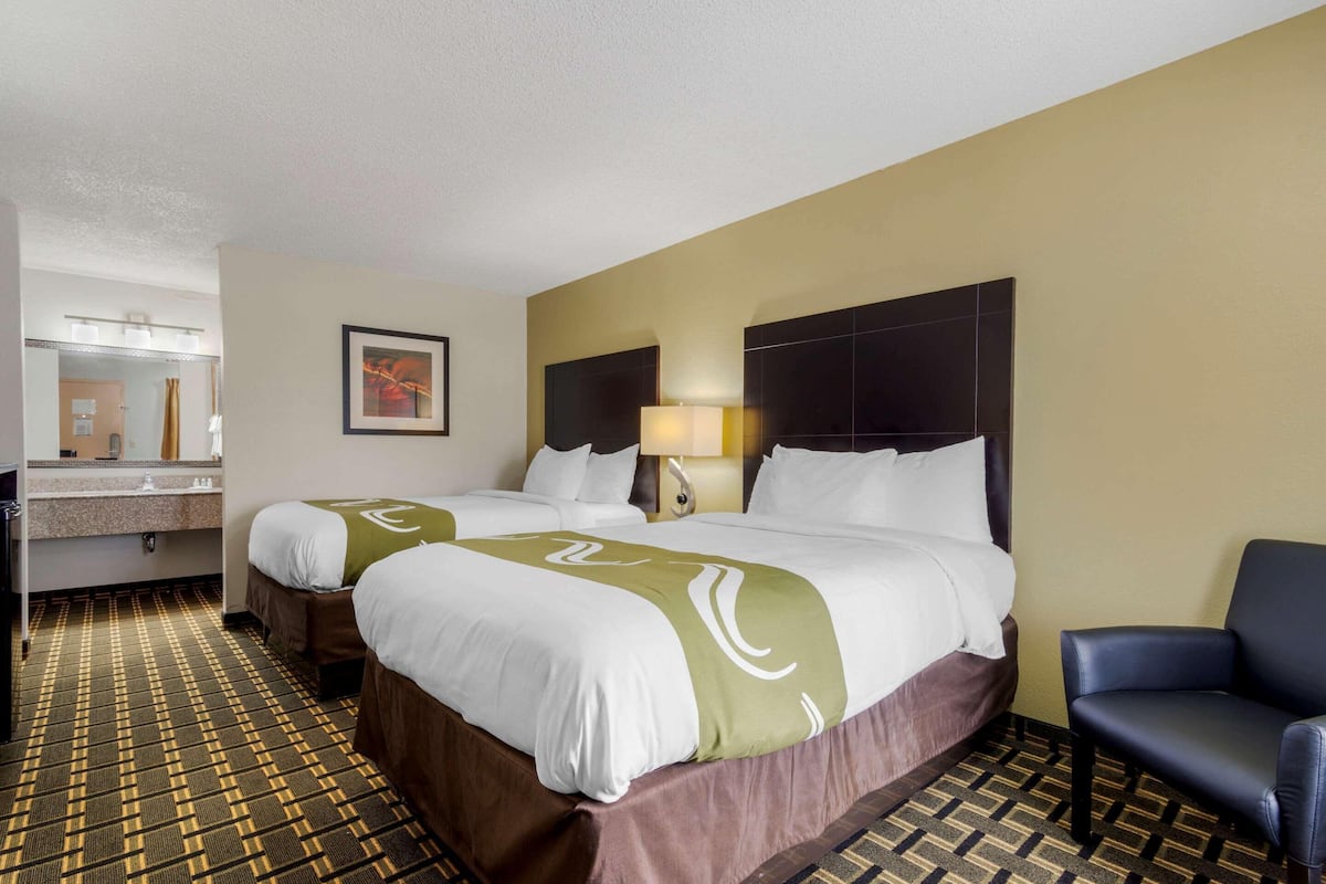 A hotel room with two beds, a bathroom, a chair, and a painting on the wall.