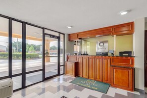 A hotel reception area with a glass door entrance, a reception desk, and a green mat with the hotel name.