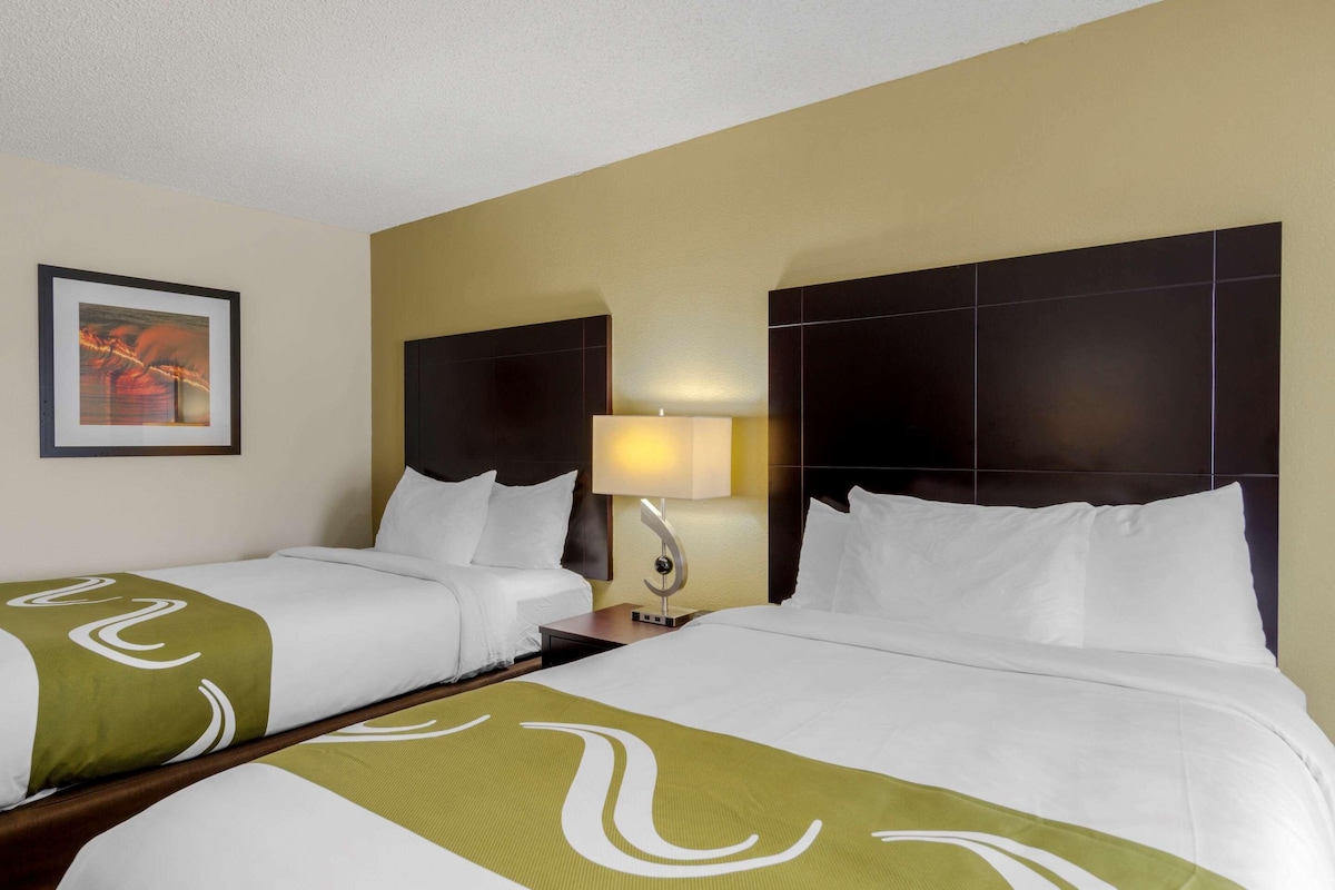 A hotel room with two beds, a nightstand, a lamp, and a framed picture on the wall.