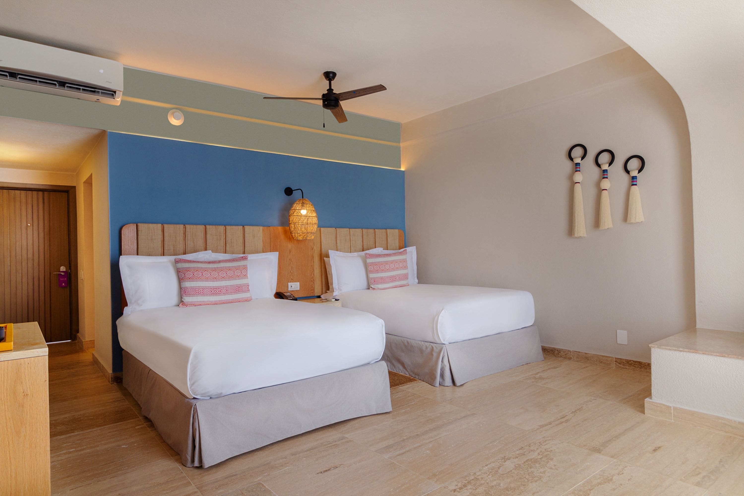 Une chambre d’hôtel avec deux lits, un ventilateur de plafond, une lampe fixée au mur et une porte en bois.