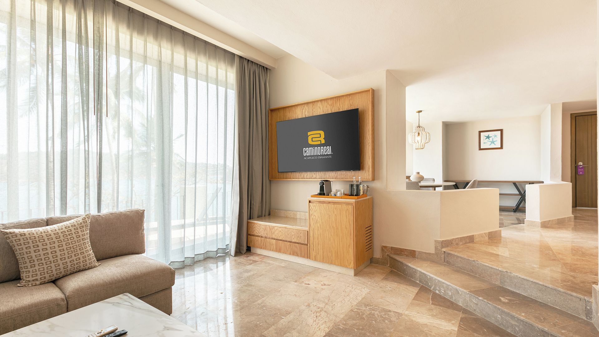 A modern hotel room with a sofa, a flat-screen TV mounted on a wooden cabinet, a dining area with a table and chairs, and a staircase leading to another level.