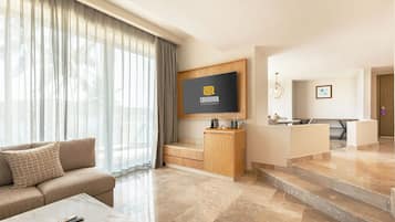 A modern hotel room with a sofa, a flat-screen TV mounted on a wooden cabinet, a dining area with a table and chairs, and a staircase leading to another level.