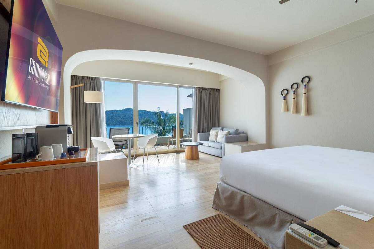 A hotel room with a large bed, a desk with a coffee maker, a sofa, and a balcony with a view of the mountains.