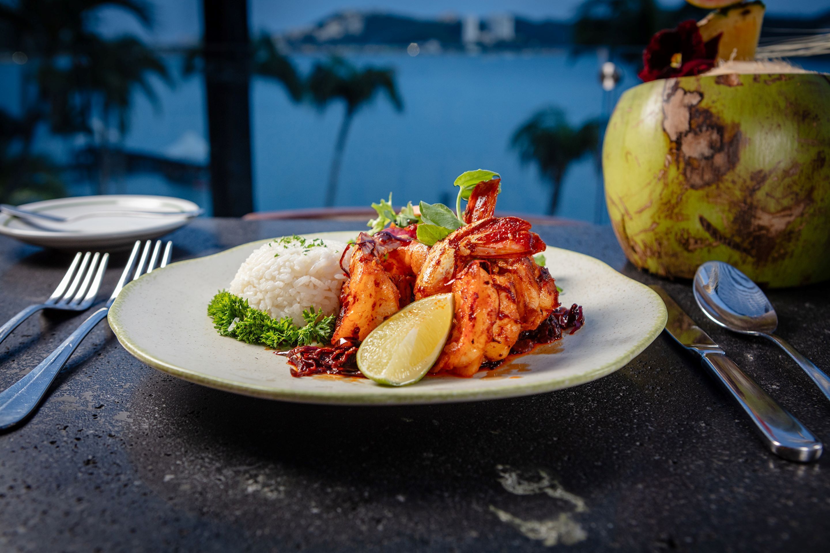 A plate of grilled chicken with rice and a lime wedge, accompanied by a coconut and utensils on a table with a view of the ocean.