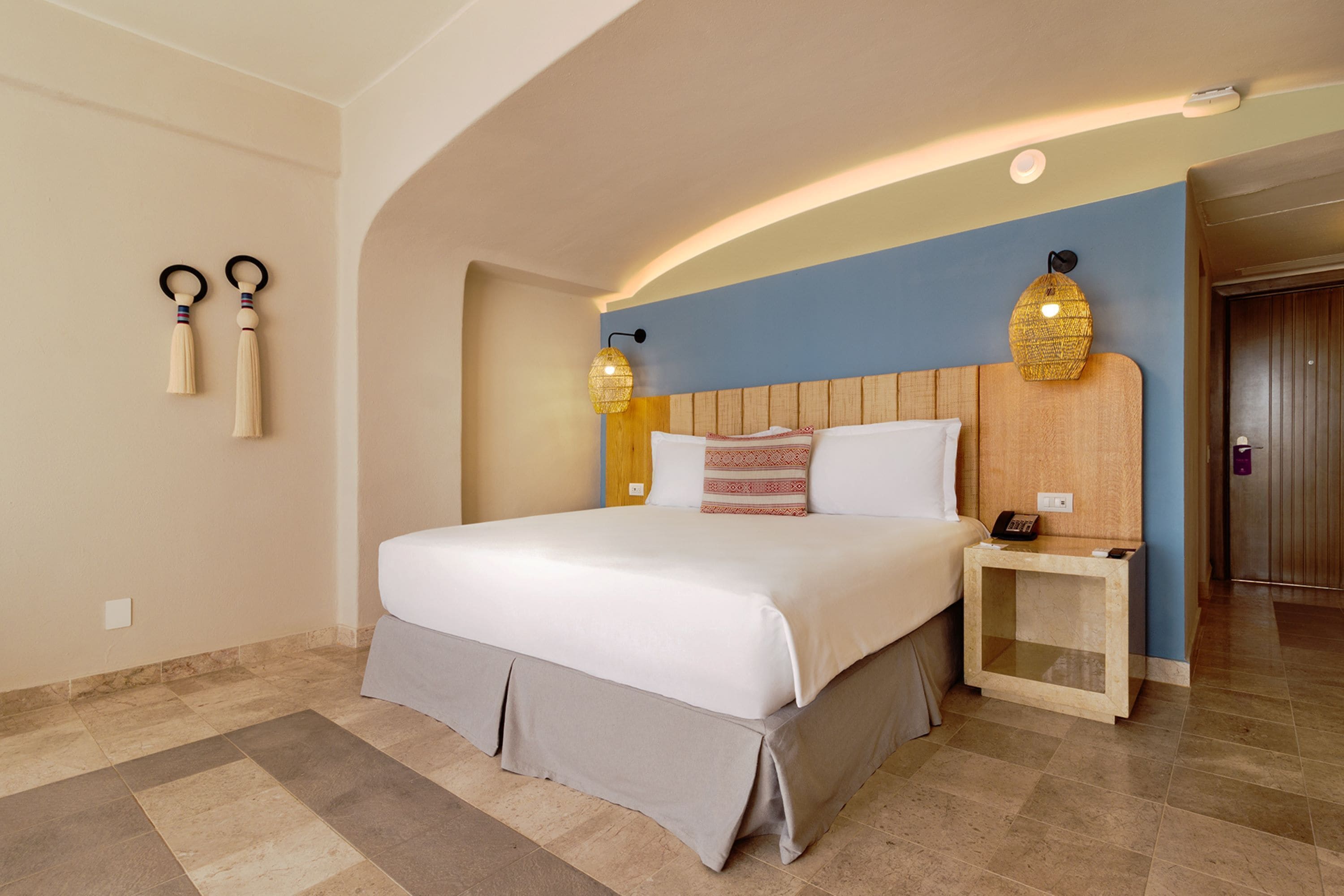 Habitación de hotel con una cama grande, cabecera de madera, dos lámparas de noche, mesitas de noche y una puerta al fondo.