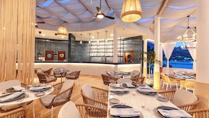 A restaurant with wicker chairs, tables set for dining, a bar area, and a view of the sea.