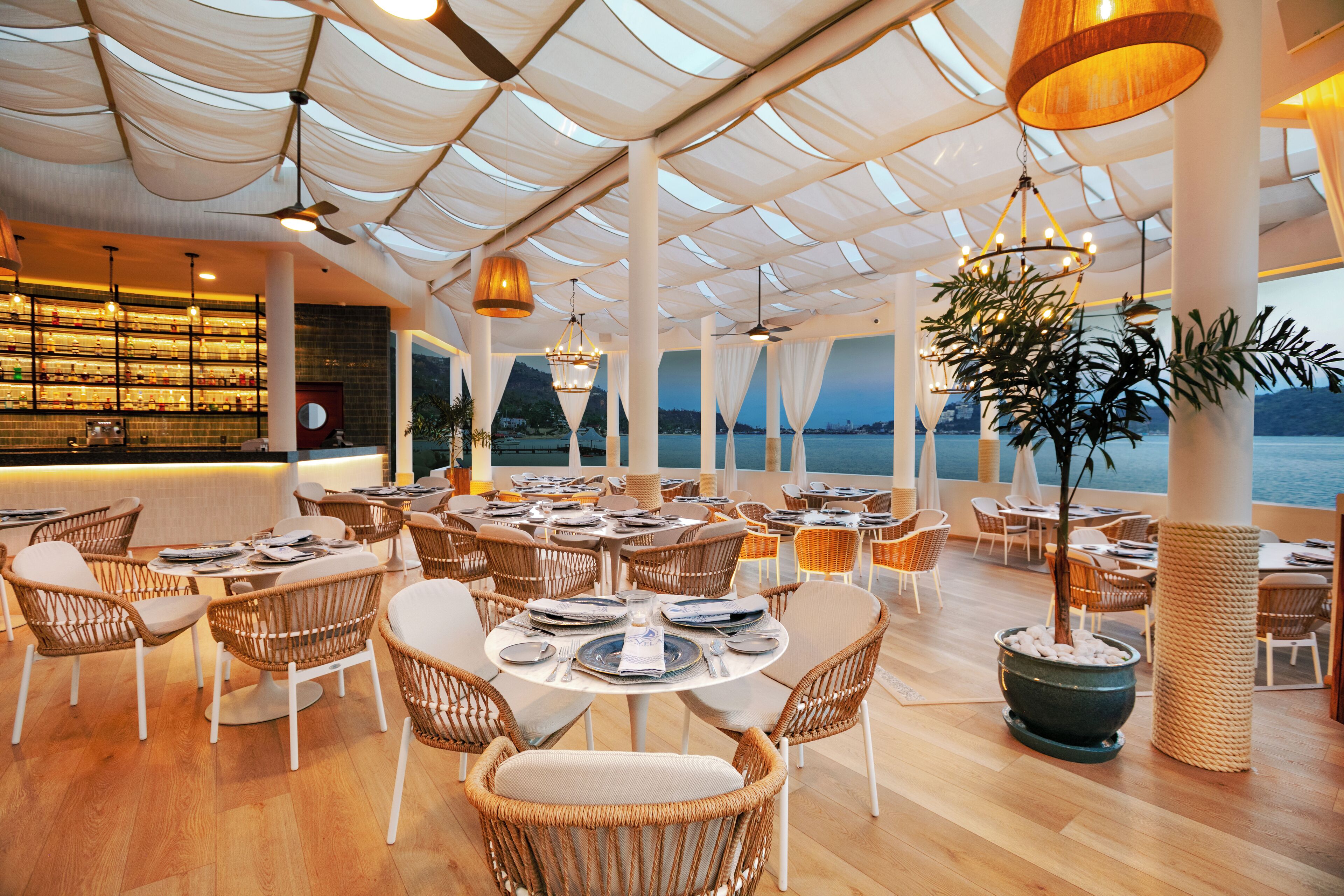 Un restaurant avec des tables rondes et des chaises en osier, un coin bar et une vue sur la mer.