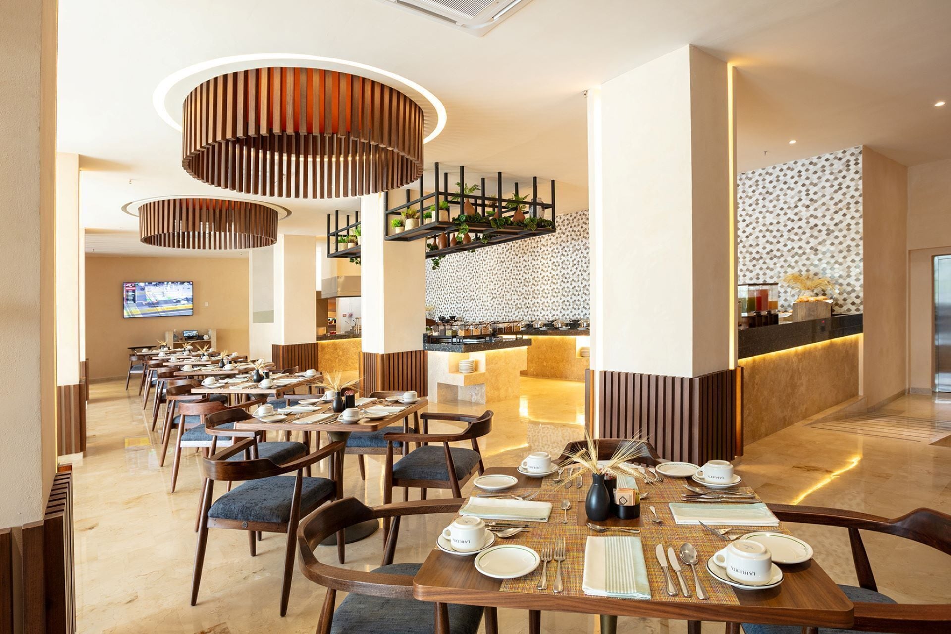 A modern hotel dining area with multiple tables set for breakfast, a bar area with a patterned backsplash, and a television screen in the background.