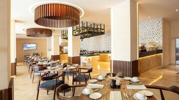 A modern hotel dining area with multiple tables set for breakfast, a bar area with a patterned backsplash, and a television screen in the background.