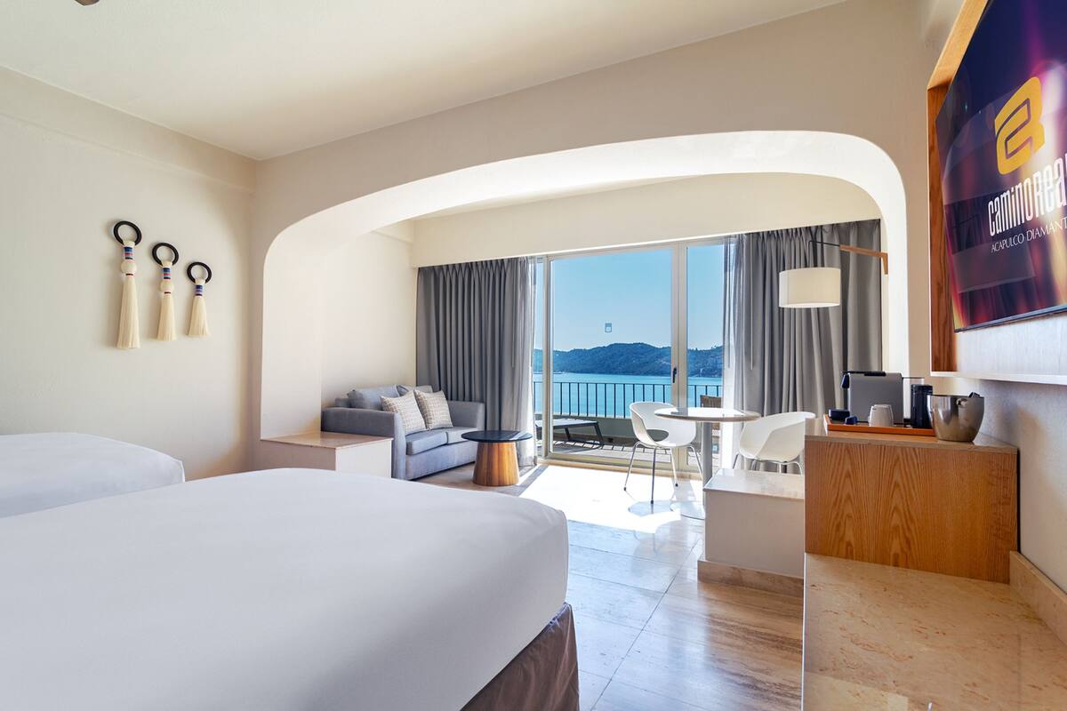 A modern hotel room with a large bed, a sofa, a dining area, and a view of the sea.