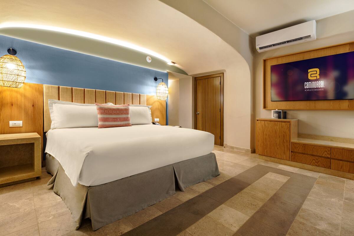 A modern bedroom with a large bed, wooden headboard, bedside lamps, a TV, and a wooden cabinet.