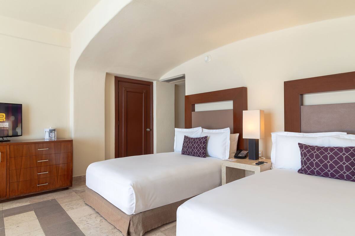 A hotel room with two beds, a wooden dresser, a television, and a nightstand with a lamp.