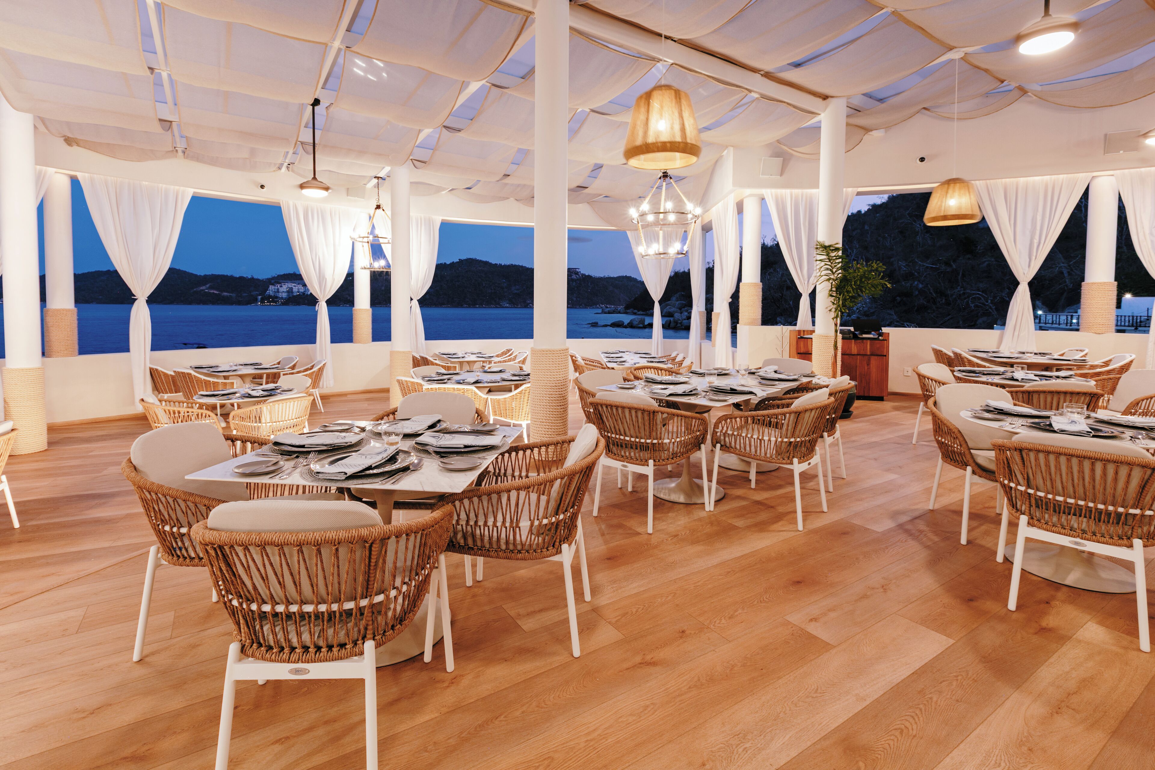 Un restaurante con sillas y mesas de mimbre, ventanas amplias con cortinas, y vista al mar.