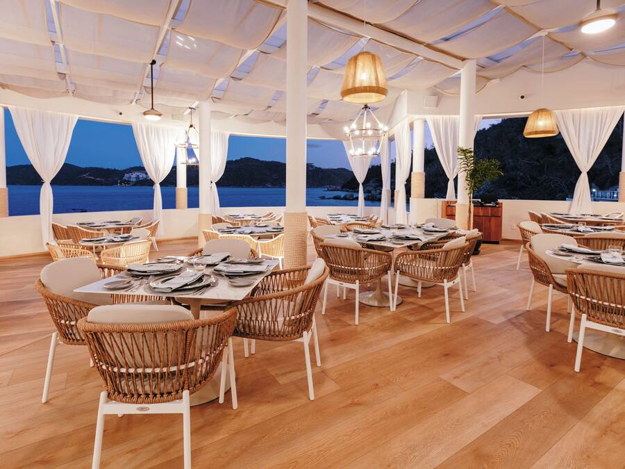 A restaurant with wicker chairs and tables, large windows with curtains, and a view of the sea.
