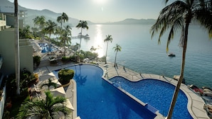A swimming pool with a view of the sea, surrounded by palm trees and a building with a poolside area.