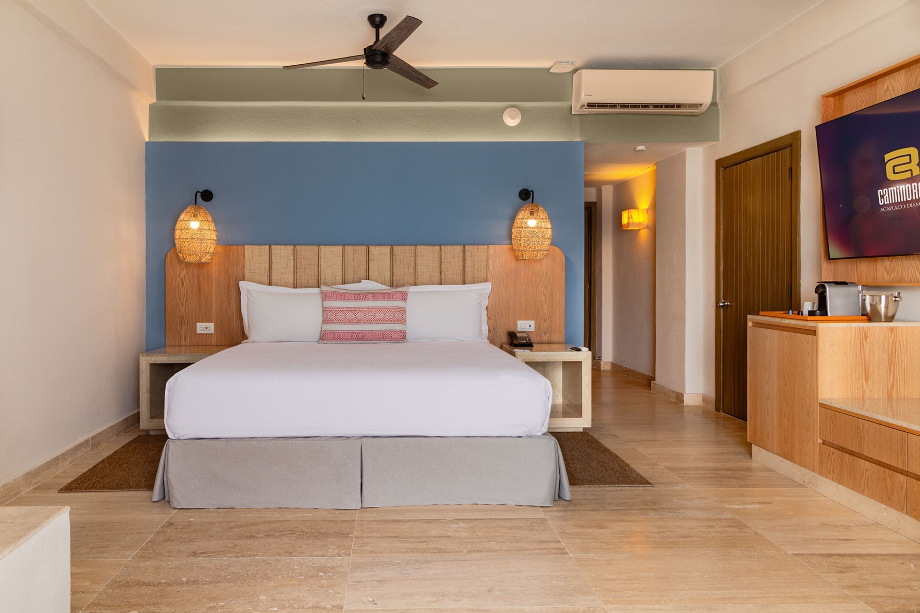 Ein modernes Hotelzimmer mit einem großen Bett, einem Holzkopfteil, zwei Nachttischlampen, einem Deckenventilator und einem Fernseher an der Wand.