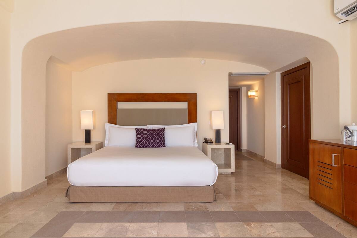 A hotel room with a large bed, two bedside tables with lamps, a wooden headboard, a wardrobe, and a hallway with a door.