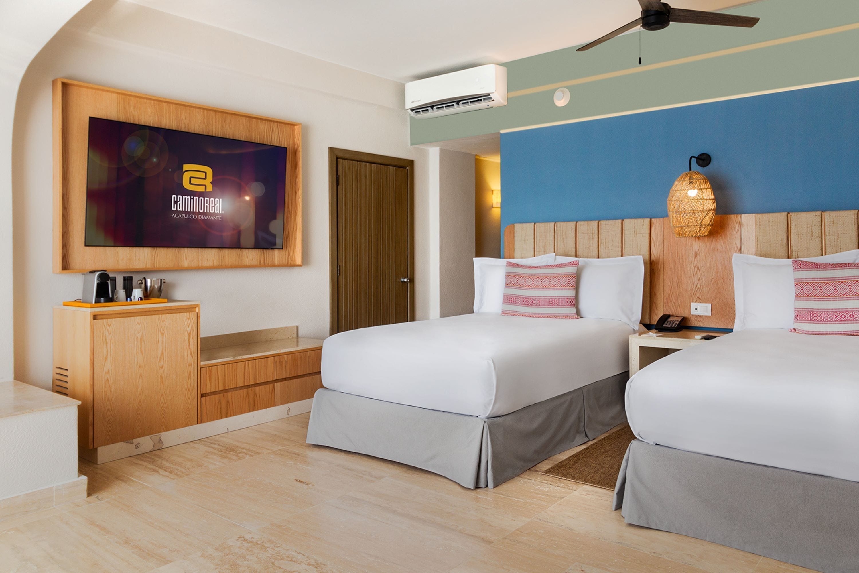 A hotel room with two beds, a TV mounted on the wall, a wooden cabinet, and an air conditioning unit.