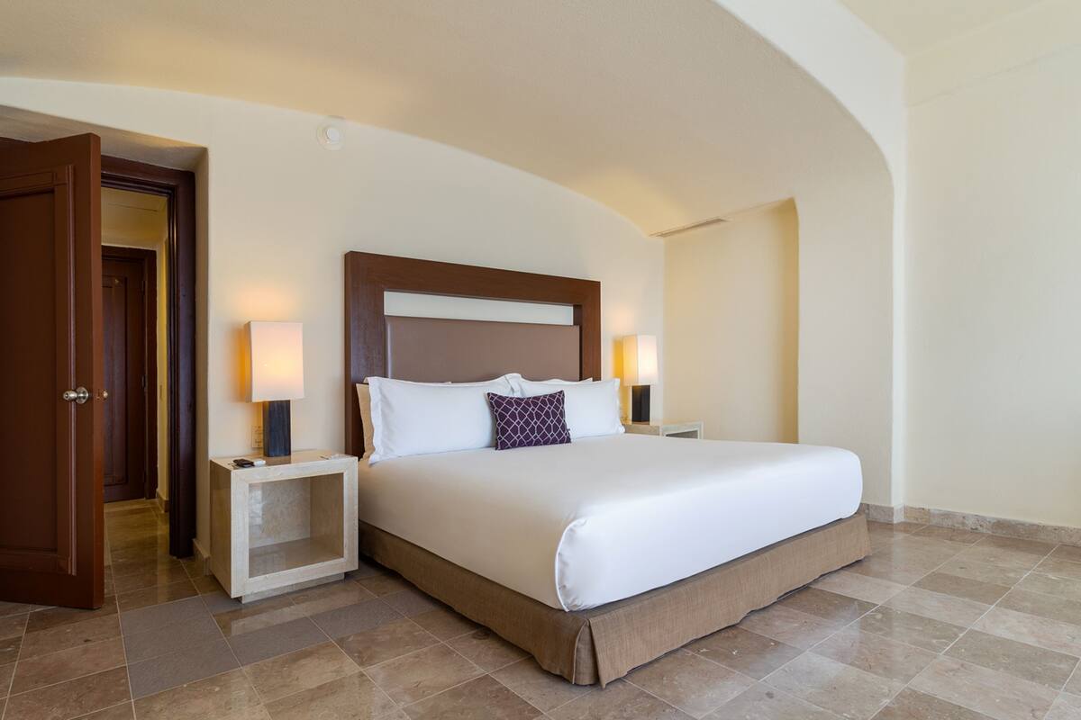 A hotel room with a large bed, two bedside tables with lamps, a wooden door, and a tiled floor.