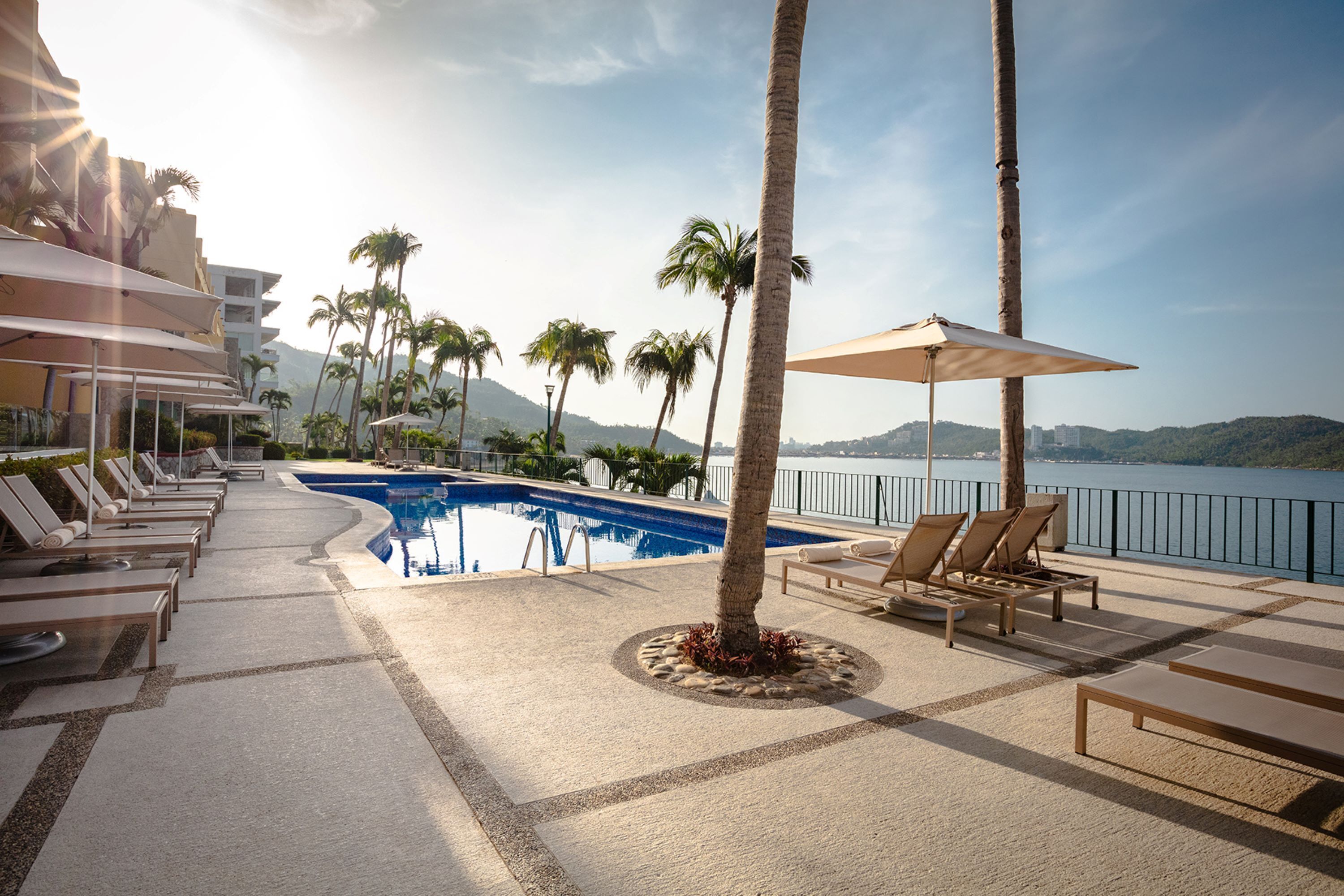 A poolside area with lounge chairs, a swimming pool, palm trees, and a view of the water.