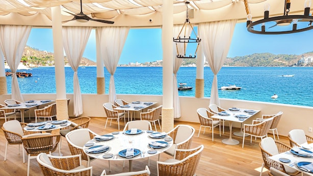 A seaside restaurant with tables and chairs arranged for dining, overlooking a clear blue sea with boats.