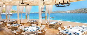 Un restaurante frente al mar con mesas y sillas dispuestas para comer, con vista a un mar azul y despejado donde se ven barcos.