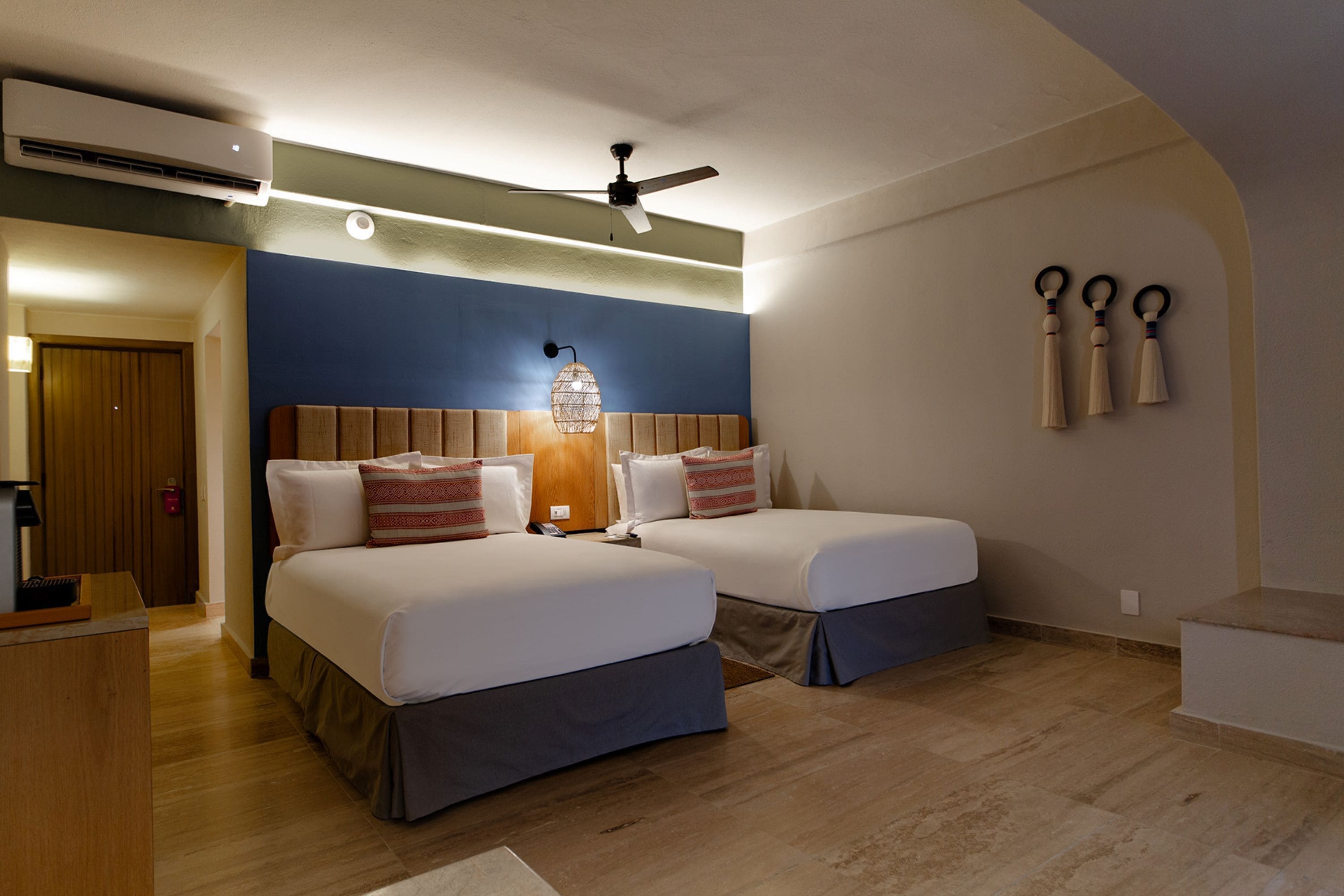 A hotel room with two beds, a ceiling fan, an air conditioning unit, a bedside lamp, and a wall-mounted telephone.