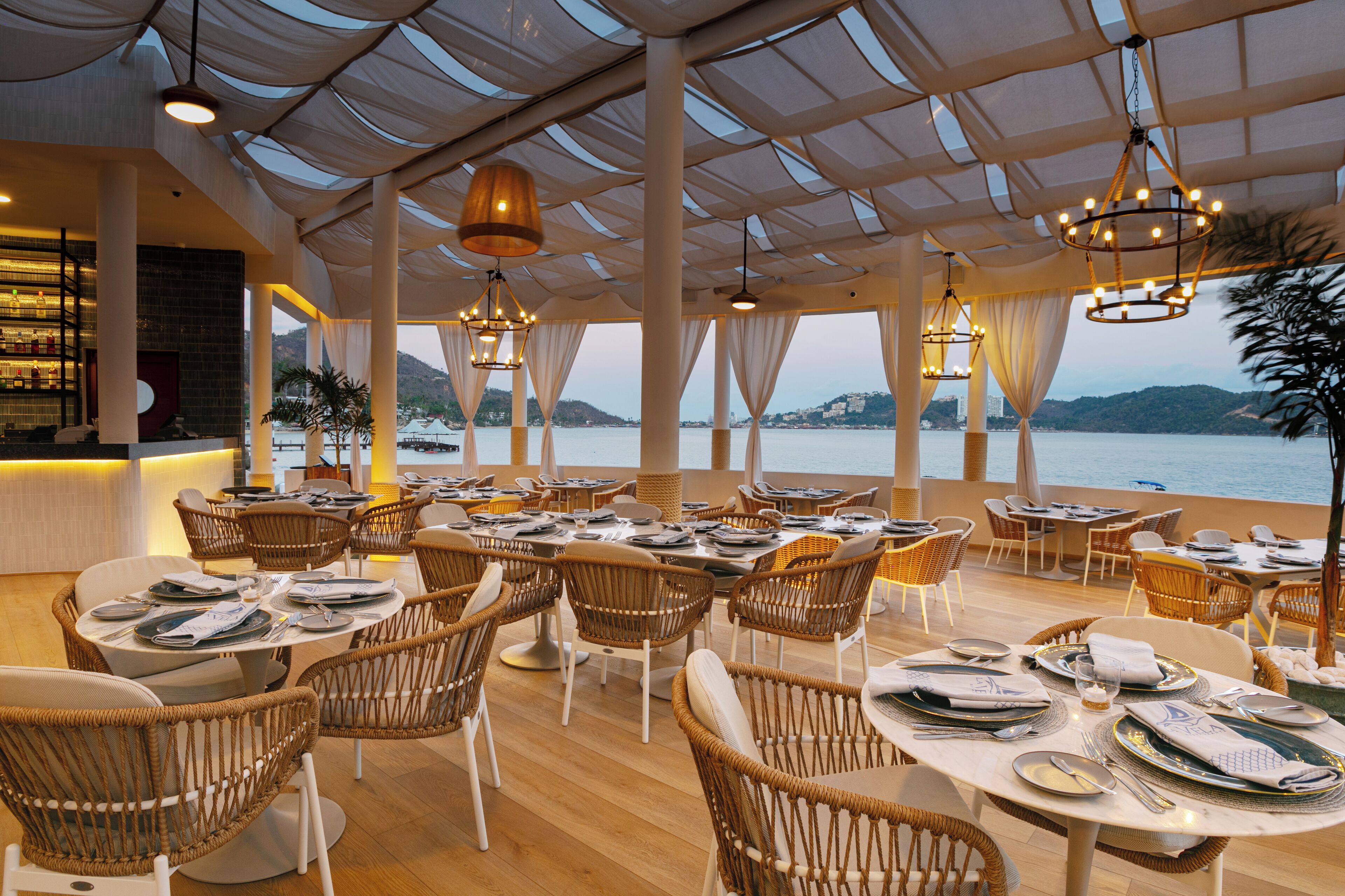 A restaurant with wicker chairs and tables set for dining, large windows offering a view of the sea, and hanging pendant lights.