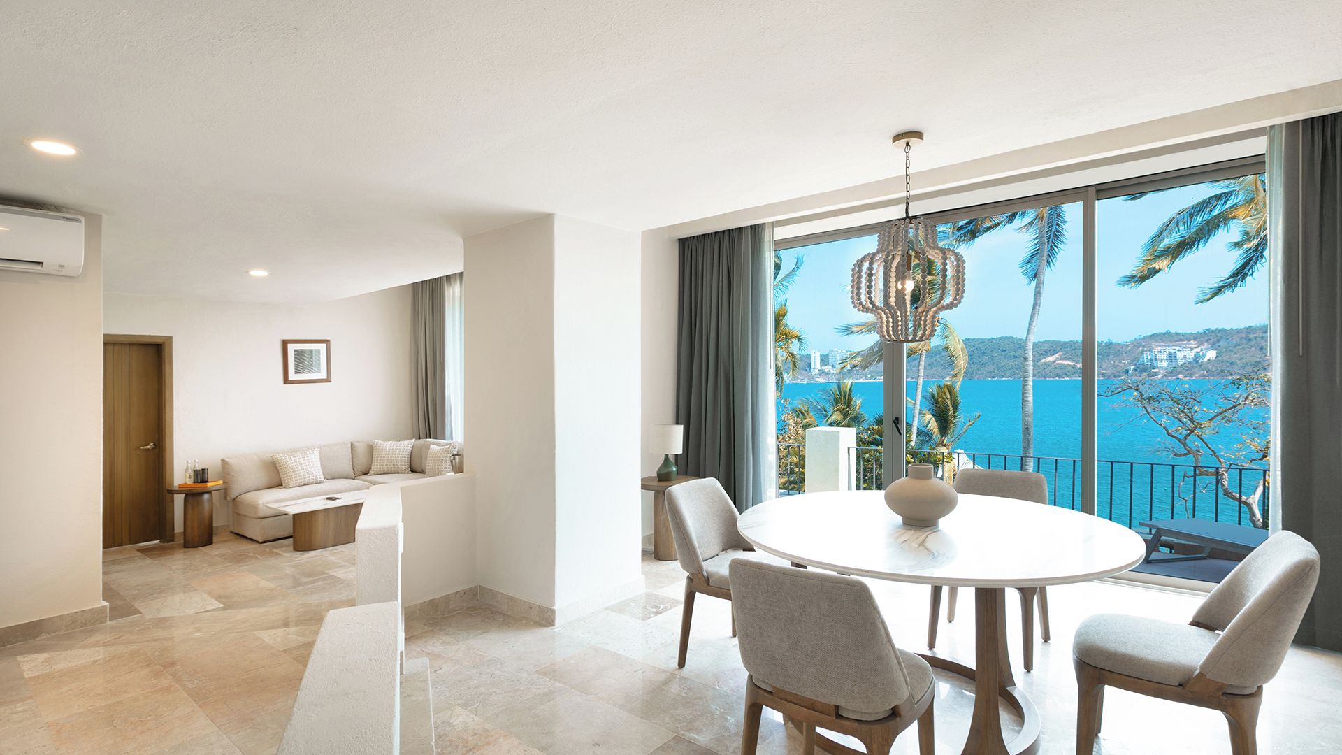 A modern living room with a round dining table, comfortable seating, and a large window offering a scenic view of the sea.