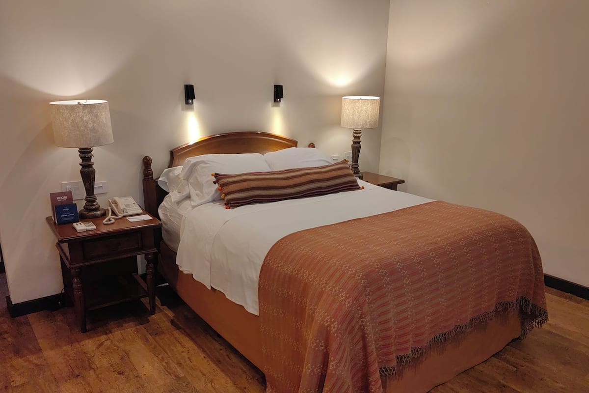 Habitación de hotel con cama de madera, ropa de cama blanca, manta marrón, dos lámparas de noche, mesita de noche con teléfono y cartel, y piso de madera.