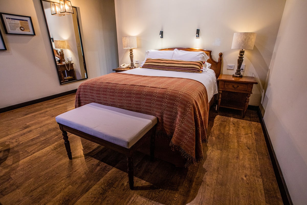 Habitación de hotel ordenada con suelo de madera, una cama grande con colcha a rayas rojas y blancas, dos mesitas de noche con lámparas, un banco al pie de la cama y un espejo grande en la pared.