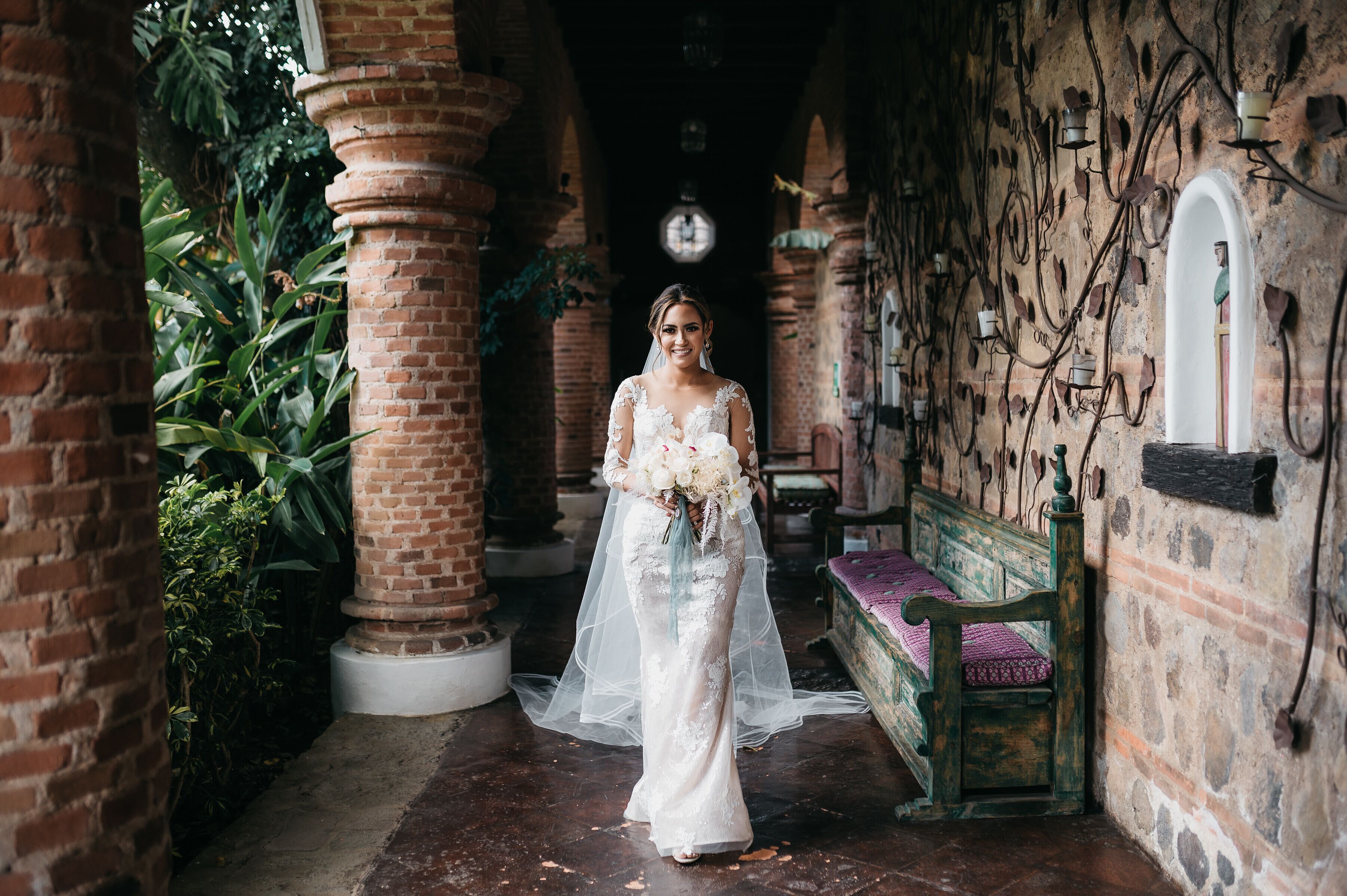 Una novia con vestido blanco parada en un pasillo con paredes de ladrillo, un banco de madera y follaje.