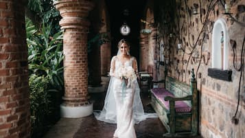 Una novia con vestido blanco parada en un pasillo con paredes de ladrillo, un banco de madera y follaje.