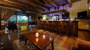 Un área de bar con vigas de madera en el techo, una barra y varios taburetes. Hay un televisor montado en la pared y una ventana que ofrece vistas al exterior.