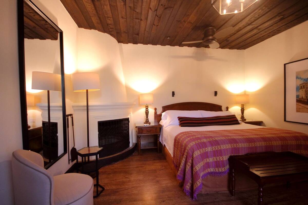 Un dormitorio acogedor con techo de madera, chimenea, cama grande con manta estampada, dos lámparas de noche y una silla.