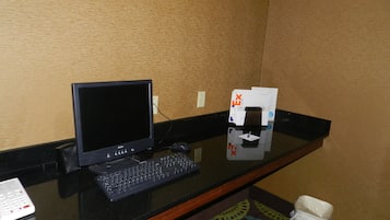 A hotel room with a desk featuring a computer, keyboard, and mouse. There is a trash bin and a small table with a pen and notepad.