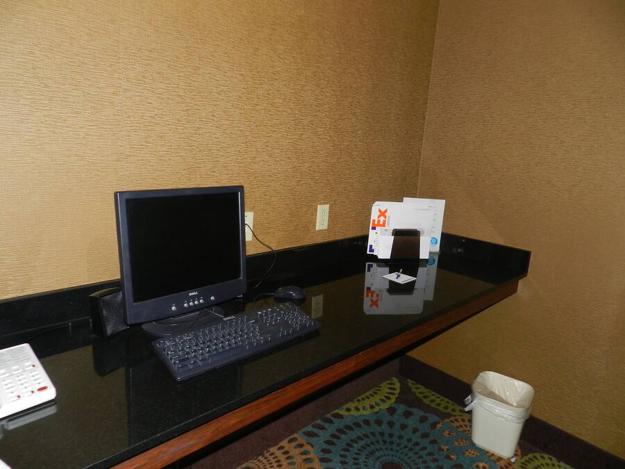 A hotel room with a desk featuring a computer, keyboard, and mouse. There is a trash bin and a small table with a pen and notepad.
