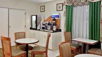 Habitación de hotel con comedor que incluye una cafetera, un refrigerador y una pequeña barra de cocina. Hay cuatro sillas alrededor de una mesa redonda y una mesa rectangular pequeña.
