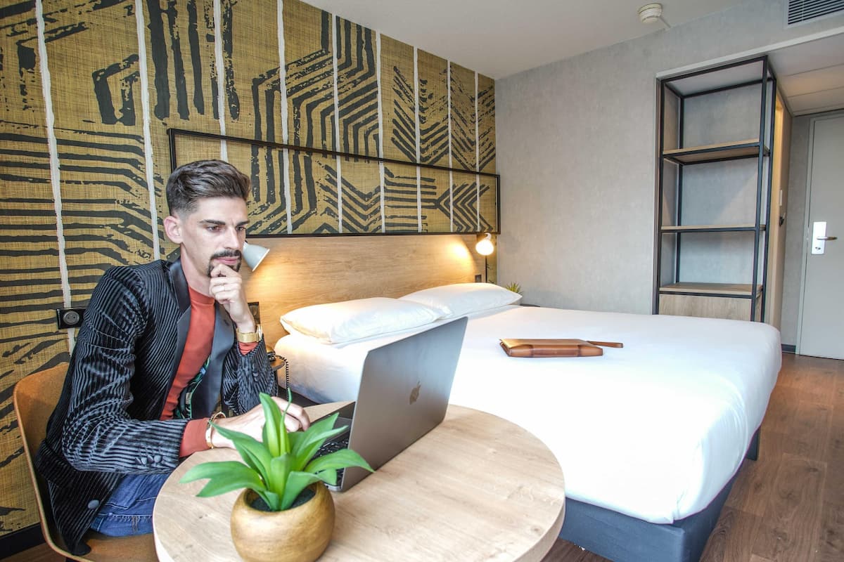 Habitación de hotel con cama, escritorio con una computadora portátil, una silla, una planta y una pared con diseño geométrico.
