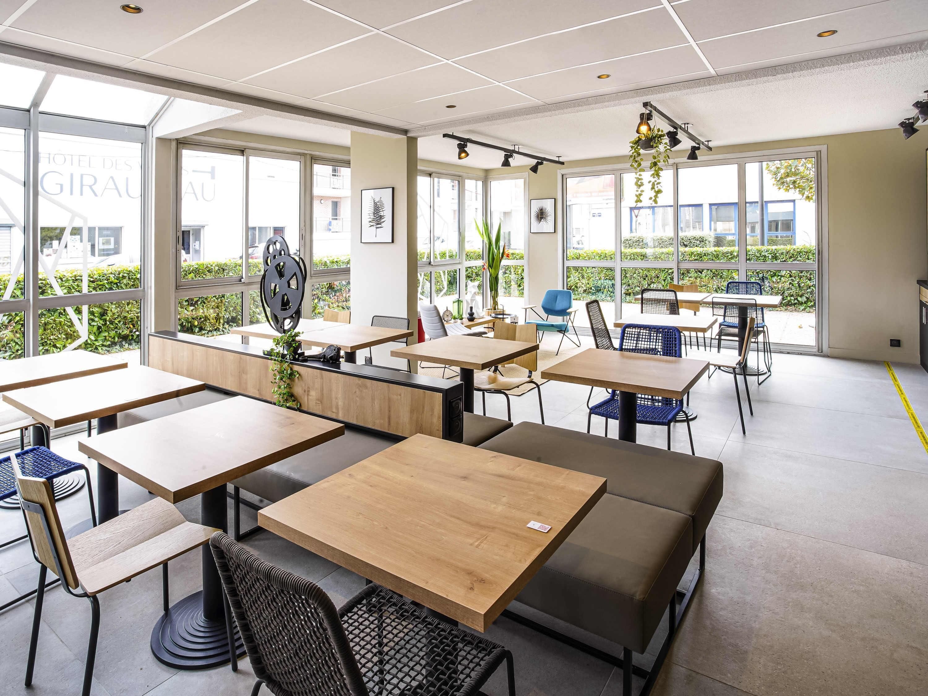 A modern dining area with multiple tables and chairs, large windows offering views of greenery, and ceiling lights.