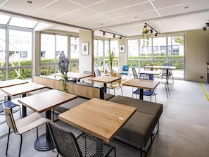 Un comedor moderno con varias mesas y sillas, ventanas amplias que ofrecen vistas a la vegetación, e iluminación en el techo.