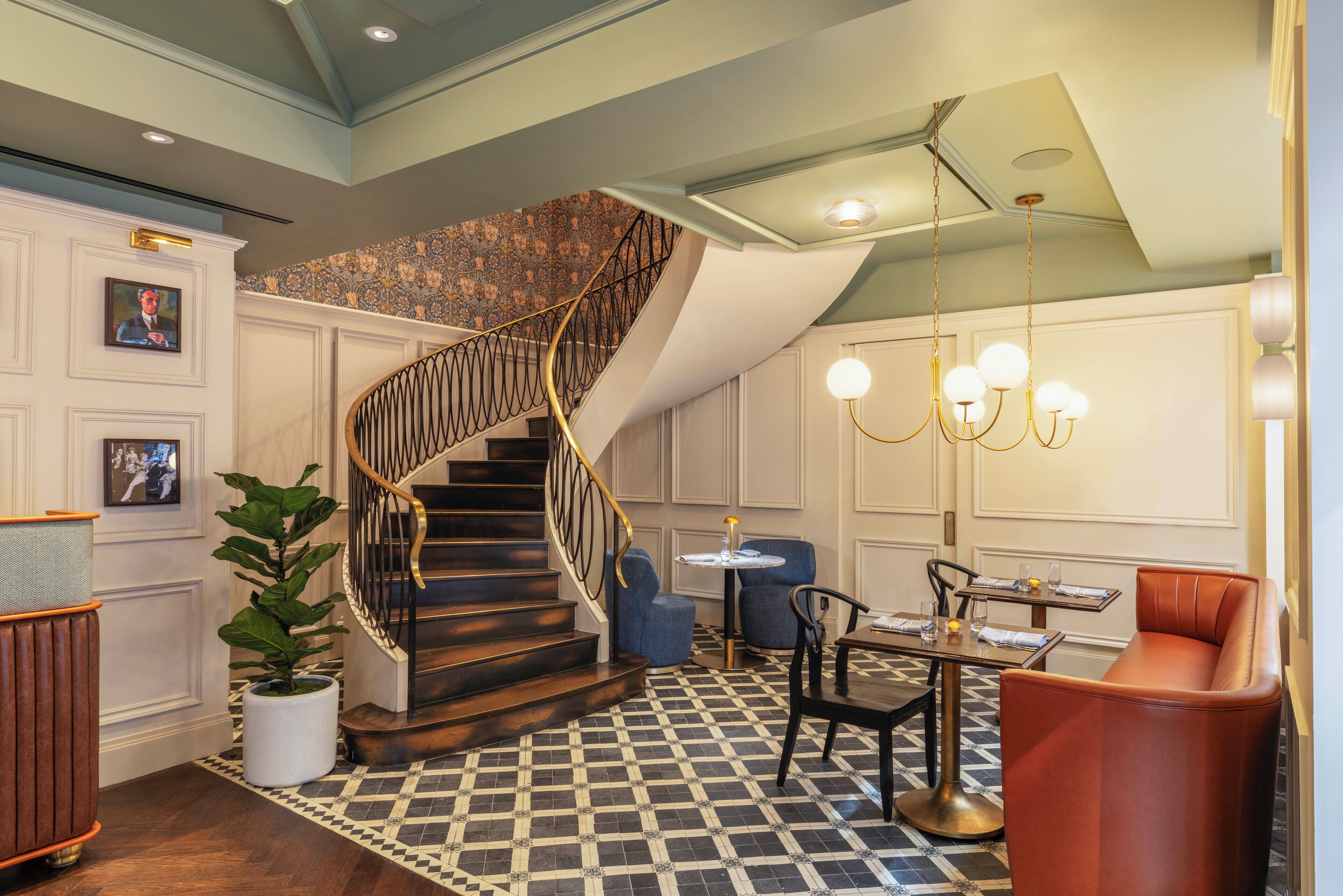 A hotel lobby with a checkered floor, a spiral staircase, a round table with chairs, and a large potted plant.