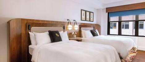 A hotel room with two beds, wooden headboards, white bedding, and a view of the outside through large windows.