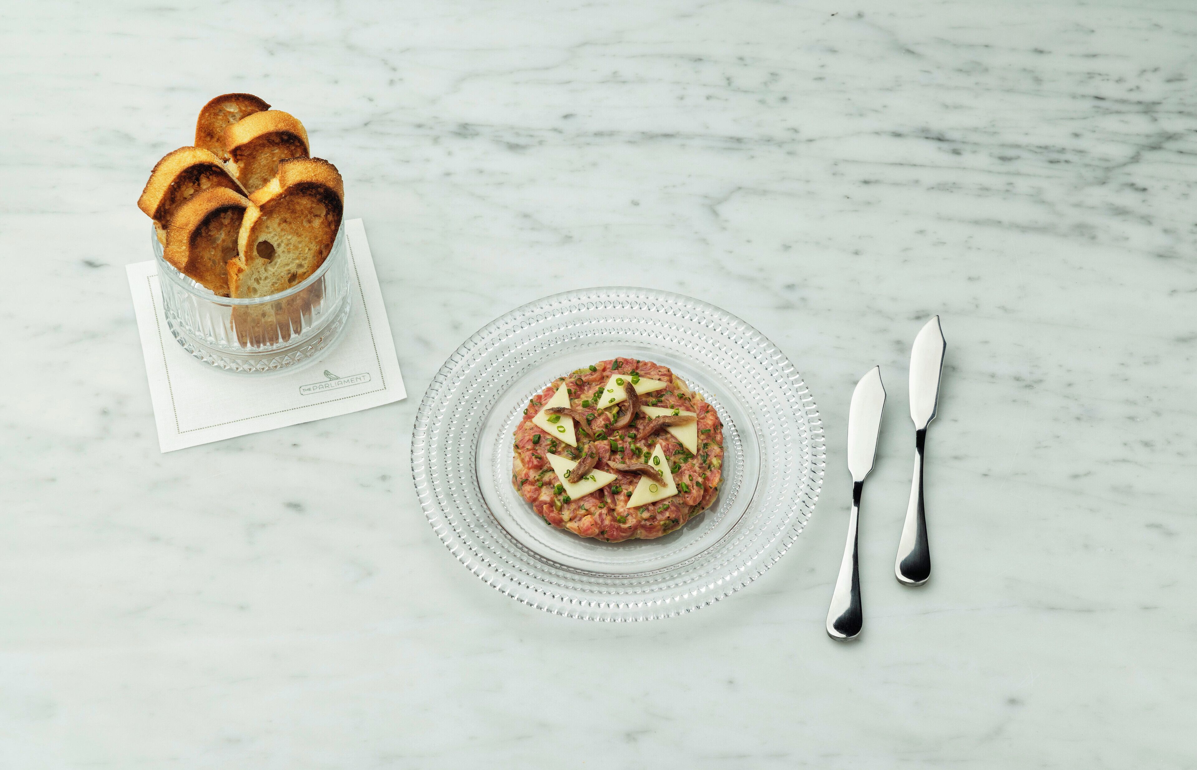 Superficie de mármol con un plato de vidrio que contiene una hamburguesa con queso y cebollines, un vaso de pan y una servilleta con un logotipo.