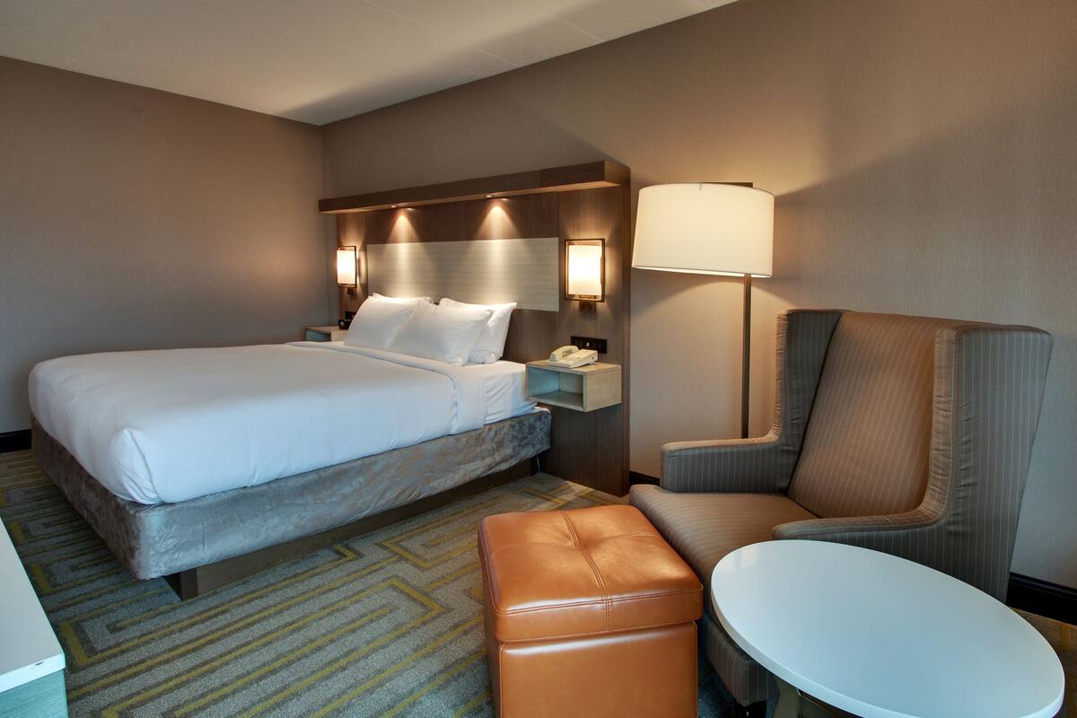 Une chambre d’hôtel avec un grand lit, une table de chevet avec une lampe, une chaise et un petit pouf.